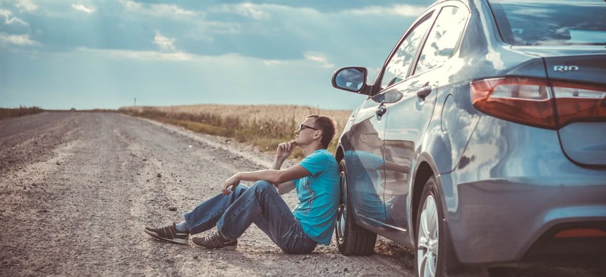 Man sitting by a car on a remote gravel road under a cloudy sky, deep in thought.